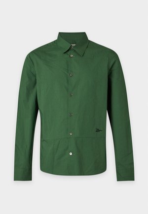Jeune homme debout portant une chemise verte à manches longues boutonnée, un pantalon marron ample et des chaussures beiges sur un fond blanc uni.