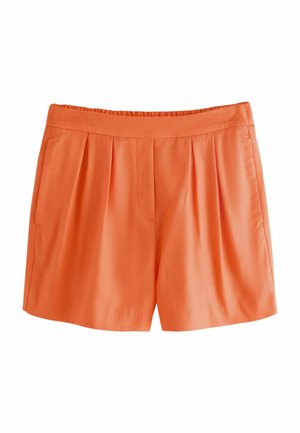 Nő, aki élénk narancssárga, testre szabott rövidnadrágot visel hátul paszpólos zsebekkel, bent áll, kezét részben az oldalzsebekben tartva.