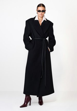 Czarny wełniany płaszcz z paskiem w talii, z dużymi klapami i długimi rękawami. Nosi się go na koszuli z paskami i zestawia z bordowymi botkami na kostkę.