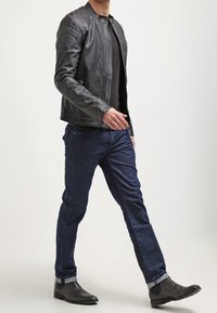 Mann läuft, trägt eine schwarze Lederjacke, ein schwarzes T-Shirt, dunkelblaue Jeans mit Umschlägen und schwarze Knöchelstiefel vor einem einfarbigen Hintergrund.