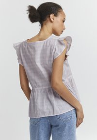 Haut en tissu léger lilas avec des manches à volants, taille froncée et texture à carreaux subtile. Conçu pour un look décontracté.