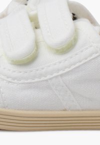 Baskets en toile blanches avec deux bandes Velcro, semelle en caoutchouc, dessus texturé et détails de couture subtils. La vue de côté met en avant le design épuré.