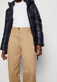 Giacca invernale blu con cappuccio, texture lucida, con polsini elastici. Pantaloni beige a vita alta con chiusura a bottone e design a gamba dritta.