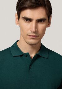 Zielona koszulka polo o teksturowanej tkaninie, z zapięciem na dwa guziki i klasycznym kołnierzykiem. Zbliżenie na odzież bez dodatkowych elementów.