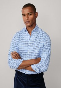 Man met gekruiste armen in een blauw-wit geruit overhemd met knopen en donkere broek tegen een effen lichte achtergrond.