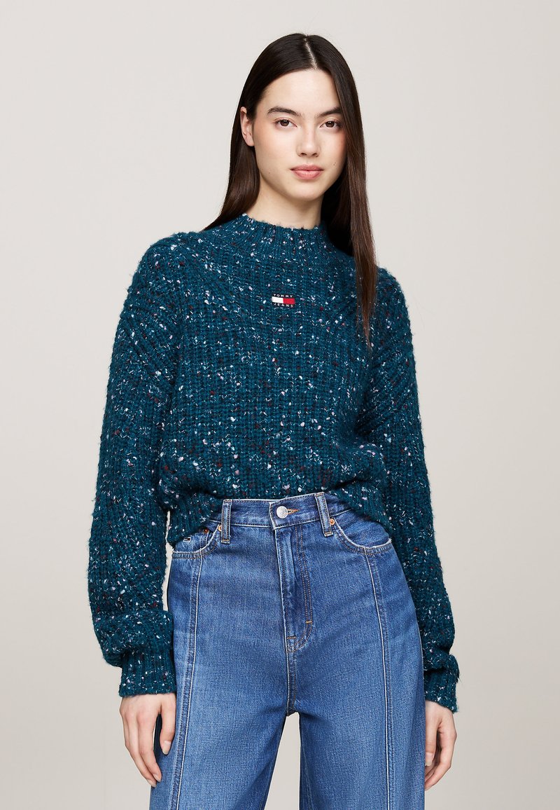 Suéter de punto verde azulado con un patrón texturizado, cuello redondo y puños acanalados, combinado con jeans azules de cintura alta que presentan bolsillos clásicos.