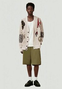 Jovem vestido com um cardigan bege estampado, t-shirt branca, calções verde-oliva, meias brancas e sapatos pretos, parado em frente a um fundo simples.