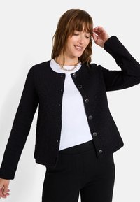 Chaqueta negra con acolchado, cuello redondo, cierre de botones en la parte delantera, mangas largas y un patrón floral texturizado. Llevada sobre una camisa blanca.
