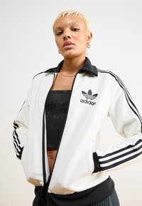White zip-up jacket featuring a black collar and three black stripes along the sleeves. The front displays the Adidas logo and trefoil.