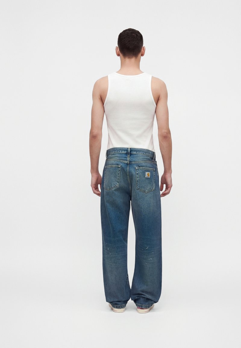Man wearing a white tank top and loose blue jeans stands facing away against a plain light gray background.