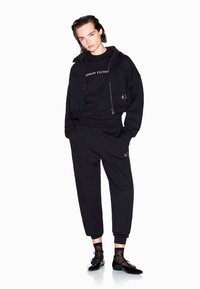 Zwart athleisure-outfit bestaande uit een cropped zip-up hoodie en bijpassende sweatpants. Het materiaal is zacht en heeft logo-accenten. Zwarte laarzen van patenterend leer maken de look compleet.