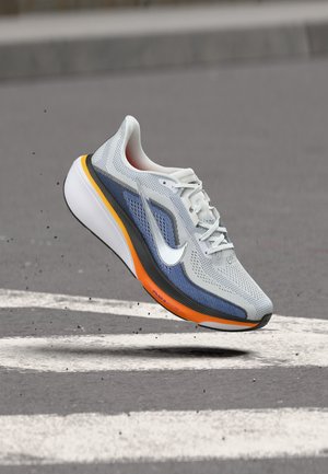Chaussure de course gris clair et bleue avec une semelle orange, suspendue au-dessus d'un bitume avec des marquages blancs au sol et de petits débris.
