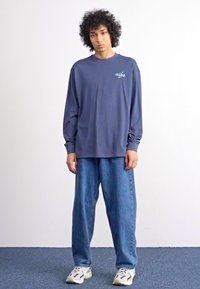Hosszú ujjú navy ing fehér Nike logóval, laza kék farmerrel és fehér sportcipővel, egy sima háttér előtt állva.