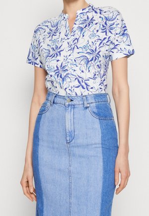Femme portant un chemisier blanc à manches courtes avec imprimé floral bleu, rentré dans une jupe en jean bleu bicolore taille haute.