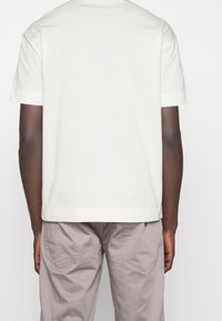 Una persona che indossa una maglietta bianca a maniche corte e pantaloni grigi chiari vista di schiena su uno sfondo bianco uniforme.