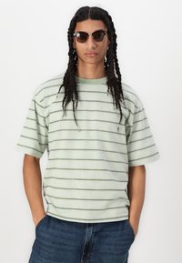 Camisa de manga corta a rayas en verde claro, con rayas horizontales en verde oscuro; combinada con pantalones cortos de mezclilla oscuros y gafas de sol.