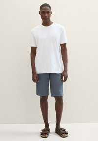 Weißes Baumwoll-T-Shirt, blaue Shorts und braune Sandalen. Das T-Shirt hat kurze Ärmel; die Shorts sind knielang und haben ein einfaches Design.