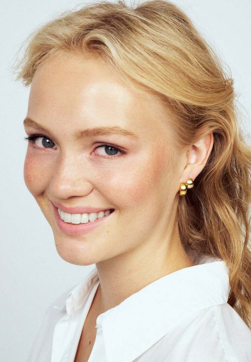 Guldtonade örhängen med en slät, rundad design och tre sammankopplade pärlor. Bakgrunden är ljus och neutral.