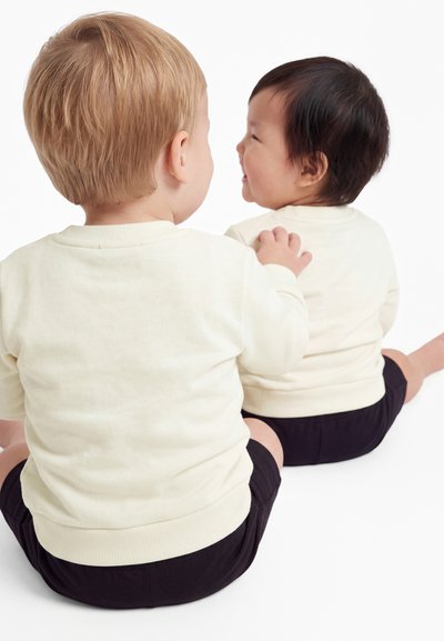Due bambini piccoli seduti per terra uno di fronte all'altro, indossano maglie color crema e pantaloncini neri, un bambino tocca la spalla dell'altro.