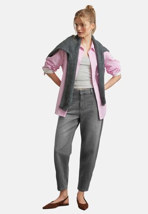 Frau mit blondem Haar im Dutt trägt ein pinkes Hemd über einem grauen Top, graue High-Waist-Jeans, einen dunkelgrauen Pullover über den Schultern drapiert und Leopardenmuster-Ballerinas.
