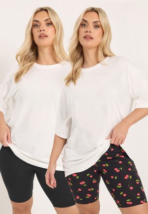 Zwei blonde Frauen tragen weiße, übergroße T-Shirts; eine trägt schwarze Radlerhosen, die andere schwarze Radlerhosen mit rotem Kirschmuster.