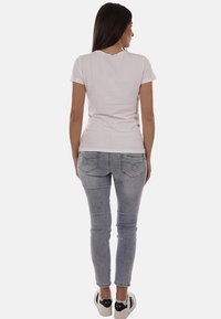 Donna in piedi con la schiena rivolta verso chi guarda, indossa una maglietta bianca semplice a maniche corte, jeans grigio chiaro e scarpe da ginnastica bianche.