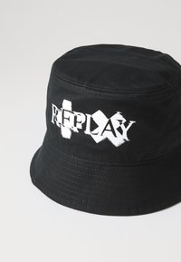Cappello a secchiello nero realizzato in cotone con logo "REPLAY" bianco a contrasto. Presenta una corona tonda e una visiera ampia e flessibile con dettagli cuciti.