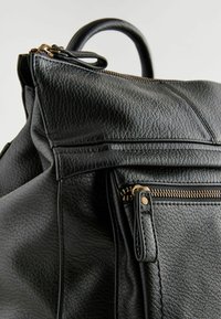 Sac à dos en cuir texturé noir avec fermeture éclair et poignée dorées, visible sur un fond clair uni.