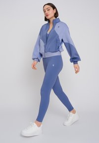 Giacca sportiva blu con design aderente, caratterizzata da un blocco colore geometrico, abbinata a leggings blu coordinati e sneakers bianche.
