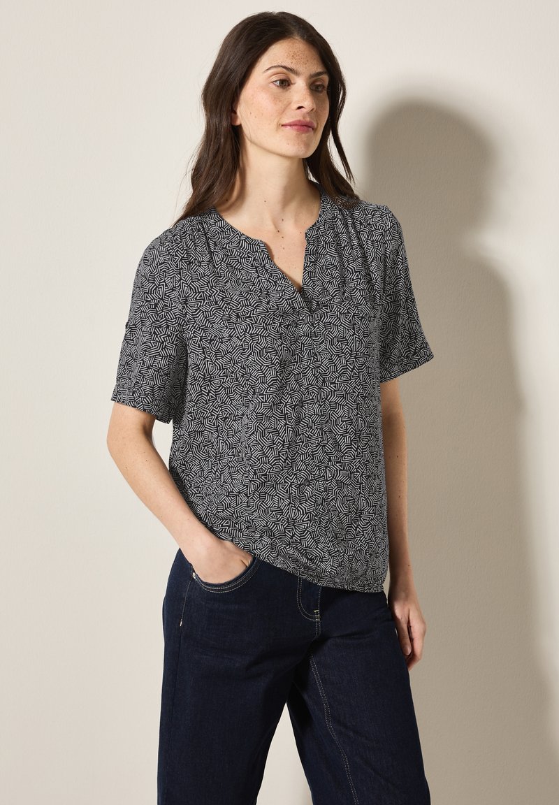 Blusa a maniche corte nera e bianca con motivo, design a V e tessuto testurizzato, indossata con pantaloni in denim blu scuro.