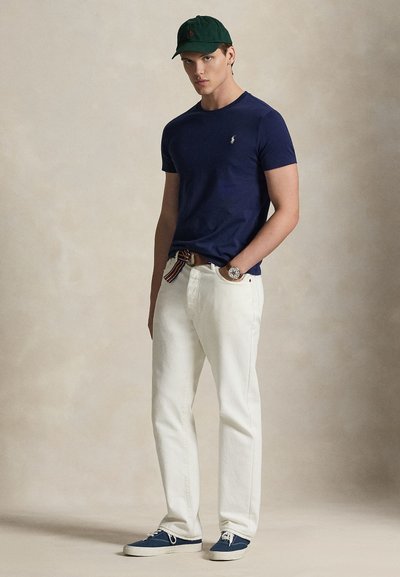 Μπλε βαμβακερό t-shirt με μικρό λογότυπο, λευκό βαμβακερό παντελόνι, πράσινο καπέλο και μπλε καμβά παπούτσια με λευκές σόλες και κορδόνια.