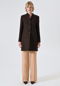 Vrouw staat gekleed in een donkerbruine, knopenblazer van lange lengte, beige wijde broek en puntige donkerbruine schoenen, tegen een effen achtergrond.