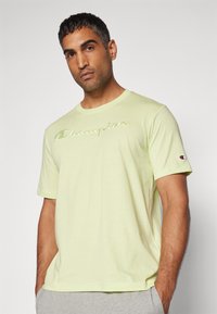 Hellgelbes Baumwoll-T-Shirt mit dem "Champion"-Logo in einem etwas dunkleren Farbton, Rundhalsausschnitt, kurzen Ärmeln und einem kleinen Logoakzent am Ärmel.