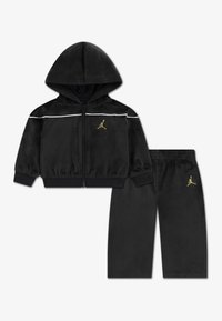 Sort velour træningsdragt med en lynlås-hoodie og matchende bukser. Har hvide striber og et gyldent logo på begge dele.