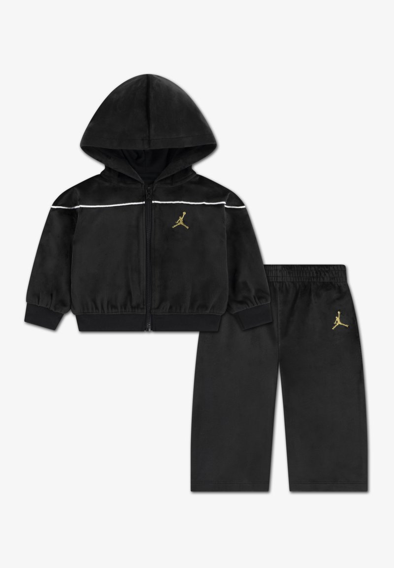 Sort velour træningsdragt med en lynlås-hoodie og matchende bukser. Har hvide striber og et gyldent logo på begge dele.