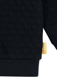 Felpa nera con motivo a pois testurizzati, polsini e orlo elasticizzati. Presenta un'etichetta gialla vicino al fondo con un logo circolare rosso.