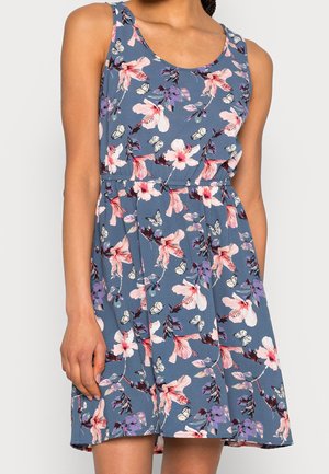 Robe bleu sans manches avec imprimé floral rose et violet et papillon blanc, portée par une personne debout, un bras détendu le long du corps.