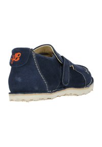 Marineblauer Wildlederschuh mit einem Klettverschluss, hellen Nähten und einem kontrastierenden orangefarbenen Logo an der Ferse. Flache Sohle mit Profil.