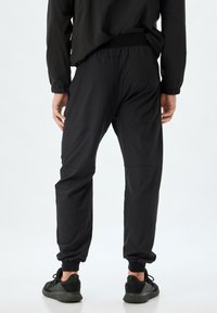 Svarta atletiska joggers med elastiska stvlar och midja, som har en slät textur och subtil sömdekor. Parat med svarta sneakers.