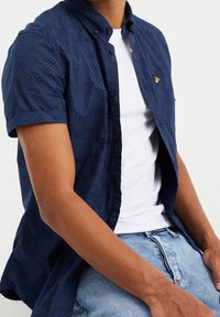 Námořnicky modrá košile s krátkým rukávem a jemným geometrickým vzorem, s límečkem na knoflíky a malou nášivkou s logem na hrudi. Nositá přes bílé tričko.