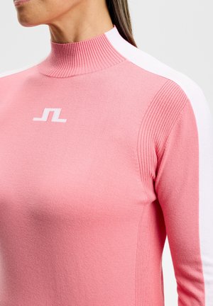 Donna che indossa un maglione rosa a collo alto con strisce bianche sulle spalle e un logo geometrico bianco sul petto.