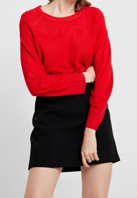 Roter, gerippter Pullover mit langen Ärmeln und lässiger Passform, kombiniert mit einem schwarzen Minirock mit seitlichen Akzenten und glatter Textur.