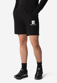 Shorts neri di cotone con vita elastica, dotati di un logo bianco e coordinate sulla coscia sinistra. Indossati con scarpe da ginnastica nere.