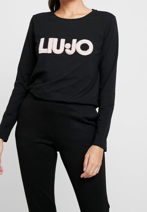 Femme portant un haut noir à manches longues avec "LIU·JO" imprimé en grandes lettres claires sur la poitrine et un pantalon noir.