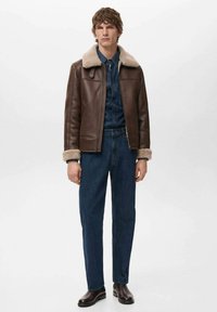 Giacca di pelle marrone con colletto in pelliccia, dotata di chiusura con zip, abbinata a una camicia di denim e jeans blu scuro. Stivali marroni completano il look.