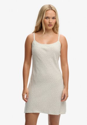 Mujer rubia con un vestido blanco sin mangas hasta la rodilla, con pequeños motivos florales, sobre un fondo blanco liso.