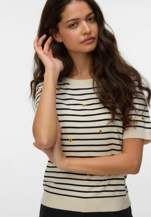 Kvinde med langt mørkt bølget hår iført en cremefarvet og sort stribet kortærmet sweater med små gule blomsterdesigns.