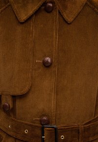 Καφέ σακάκι από κορντόνι με μεγάλα στρογγυλά καφέ κουμπιά, φαρδιά γιακά, τσέπη με καπάκι και ταιριαστή ζώνη από κορντόνι με μαύρη αγκράφα.