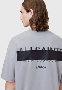 T-shirt oversized gris à manches courtes. Présente une bande noire imposante avec le texte en relief "ALLSAINTS" et "LONDON" en dessous.