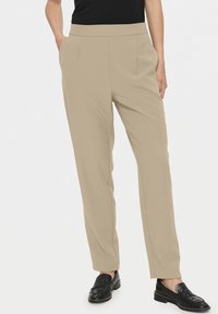 Pantalons beige ajustés au toucher doux, avec des poches latérales et une coupe droite, associés à des mocassins noirs.